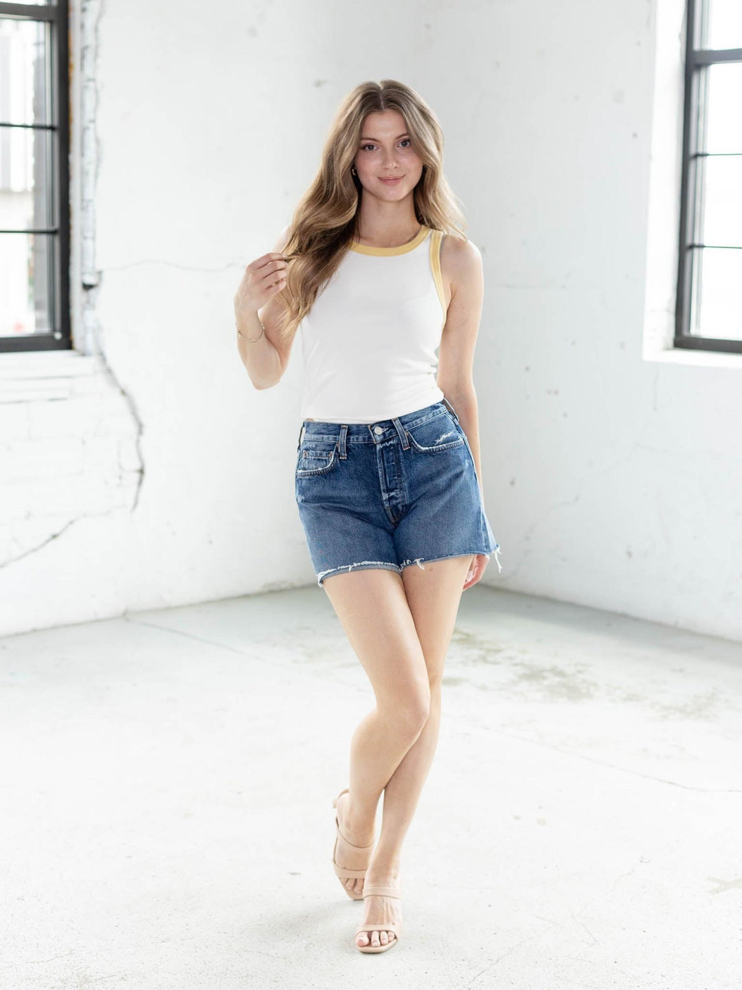 Model wearing a white ribbed cotton tank top with yellow contrast trim, blue denim shorts, and nude sandals.