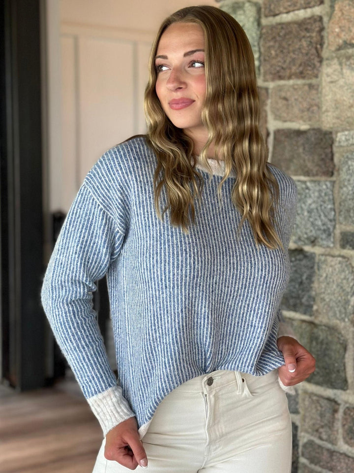 A woman wearing a light blue and white vertically striped knit sweater with ribbed cuffs, paired with cream-colored pants, standing in front of a stone wall.
