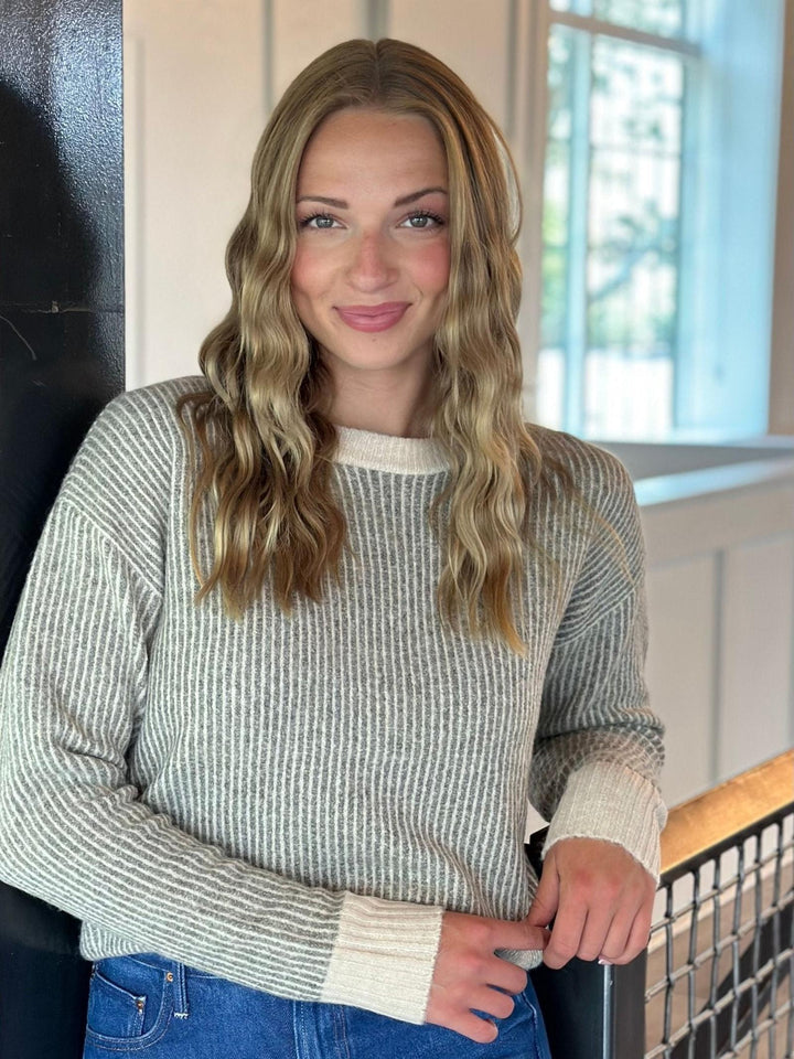 A woman with long wavy blonde hair wearing a ribbed knit crewneck sweater in a light gray and white pattern, paired with blue jeans, standing indoors near a railing.