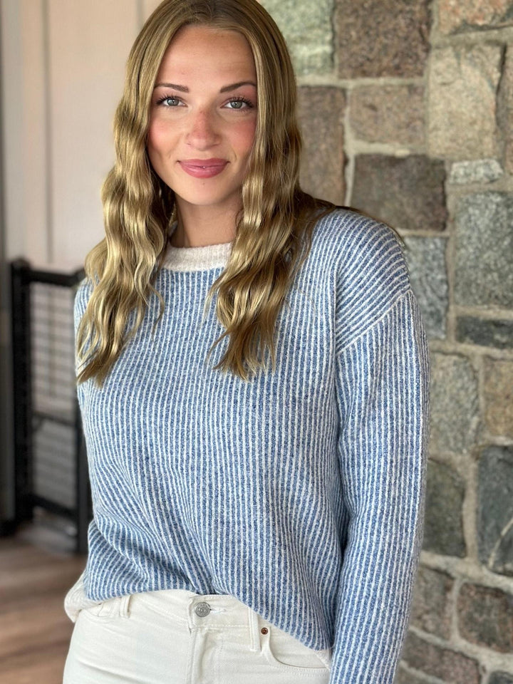 A woman with long blonde wavy hair wearing a light blue and white vertically ribbed knit sweater and white pants, standing in front of a stone wall.