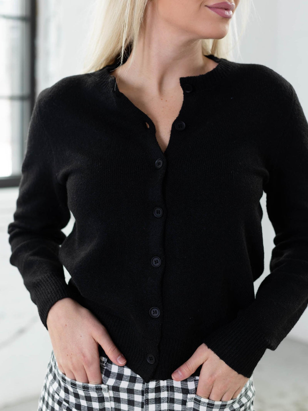 Woman wearing a black cardigan over a checkered dress in a bright room.
