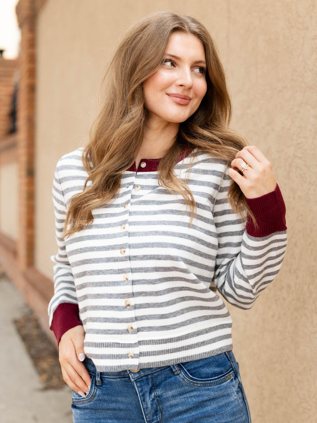 A woman wearing a gray and white striped button-up cardigan with burgundy ribbed trim and cuffs, paired with blue jeans, standing against a beige stucco wall.