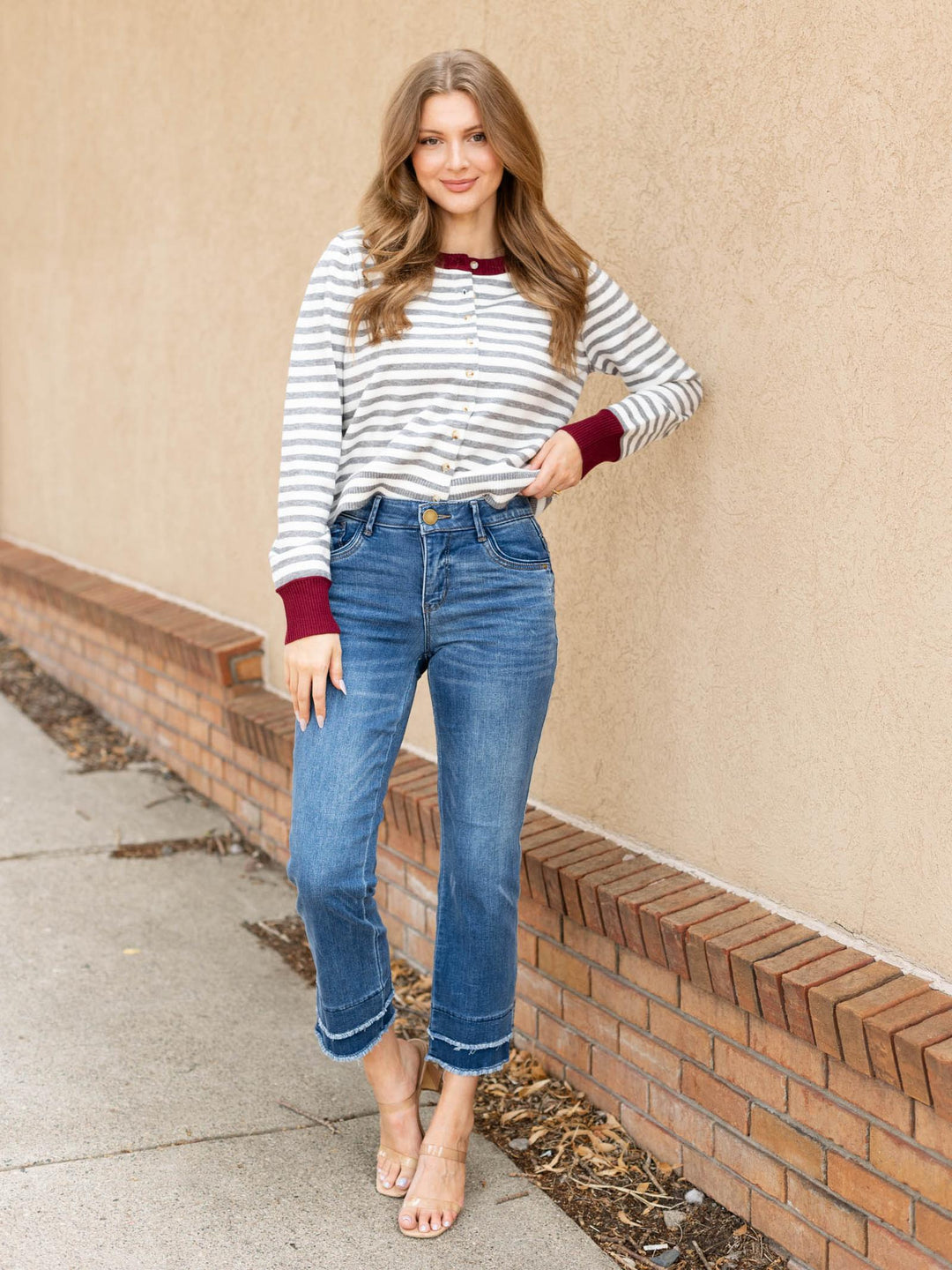 A woman stands outdoors against a beige stucco wall and brick baseboard, wearing a white long-sleeve top with thin gray horizontal stripes and dark burgundy cuffs, paired with blue cropped jeans and clear high-heel sandals.