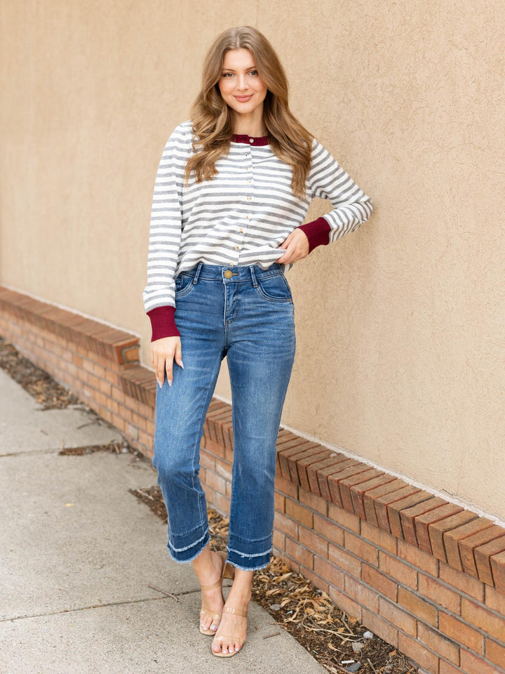 A woman stands outdoors against a beige stucco wall and brick baseboard, wearing a white long-sleeve top with thin gray horizontal stripes and dark burgundy cuffs, paired with blue cropped jeans and clear high-heel sandals.