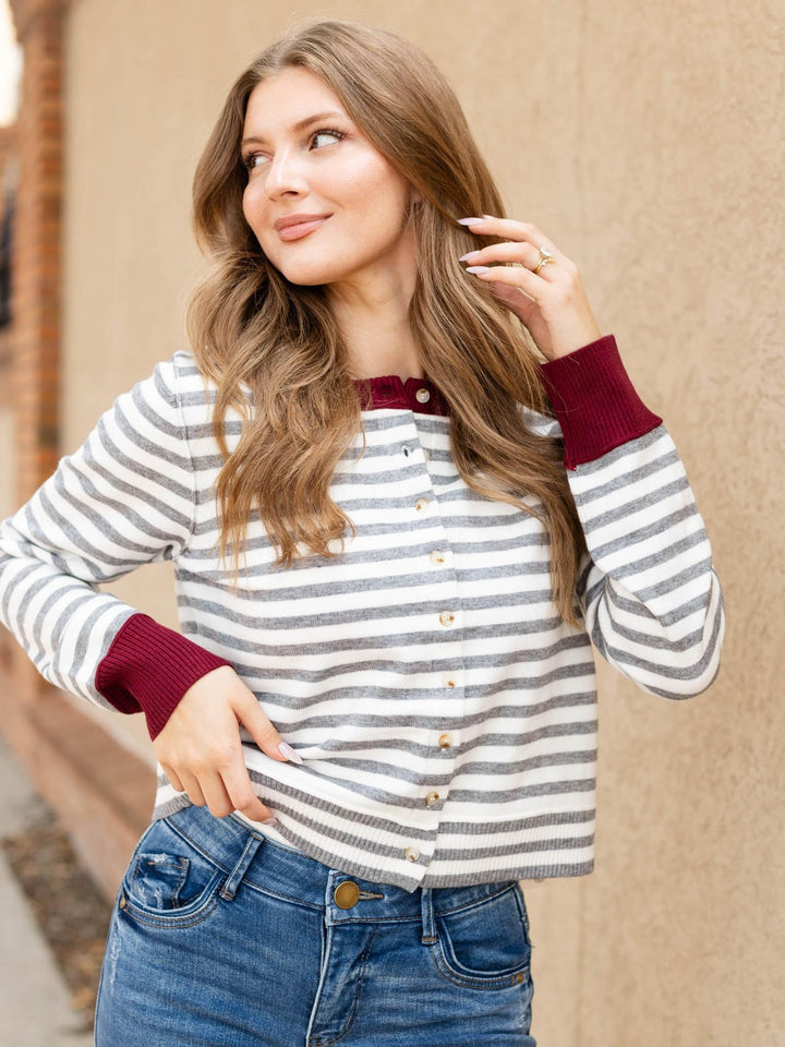 A woman wearing a gray and white striped long-sleeve cardigan with burgundy ribbed trim and buttons, paired with blue jeans, standing against a beige wall.