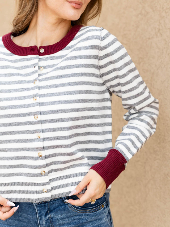 A woman wearing a long-sleeve cardigan with horizontal gray and white stripes, featuring a deep burgundy ribbed crew neck, button closure, and matching burgundy ribbed cuffs, paired with blue jeans.