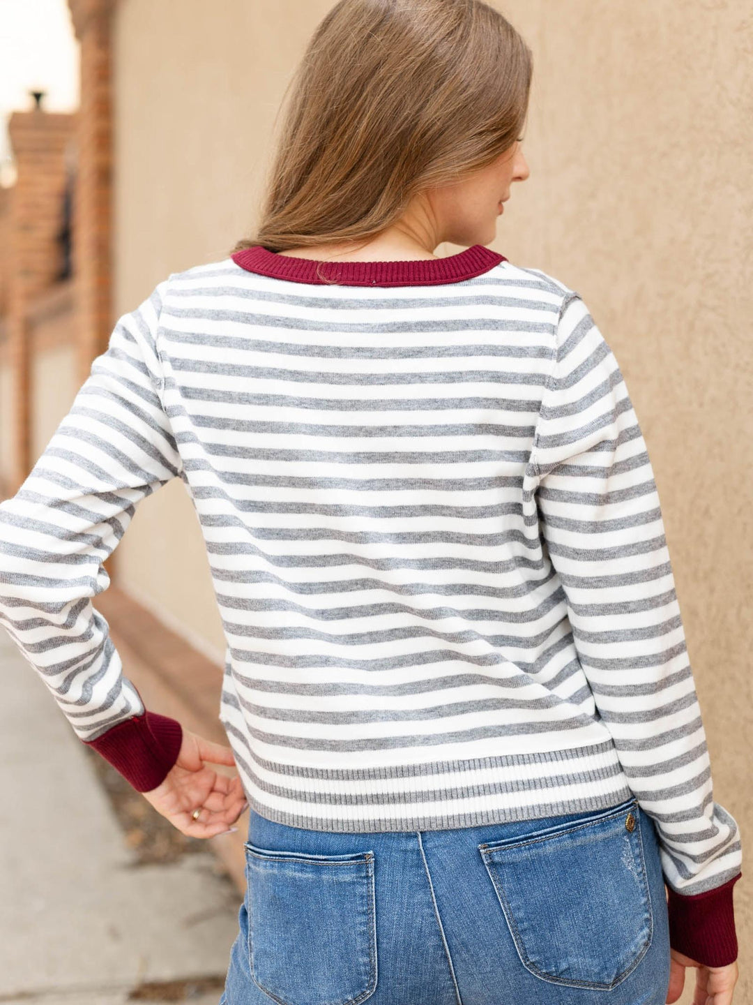 A woman shown from the back wearing a gray and white horizontally striped long-sleeve sweater with burgundy ribbed trim at the crew neck, cuffs, and hem, paired with blue jeans.