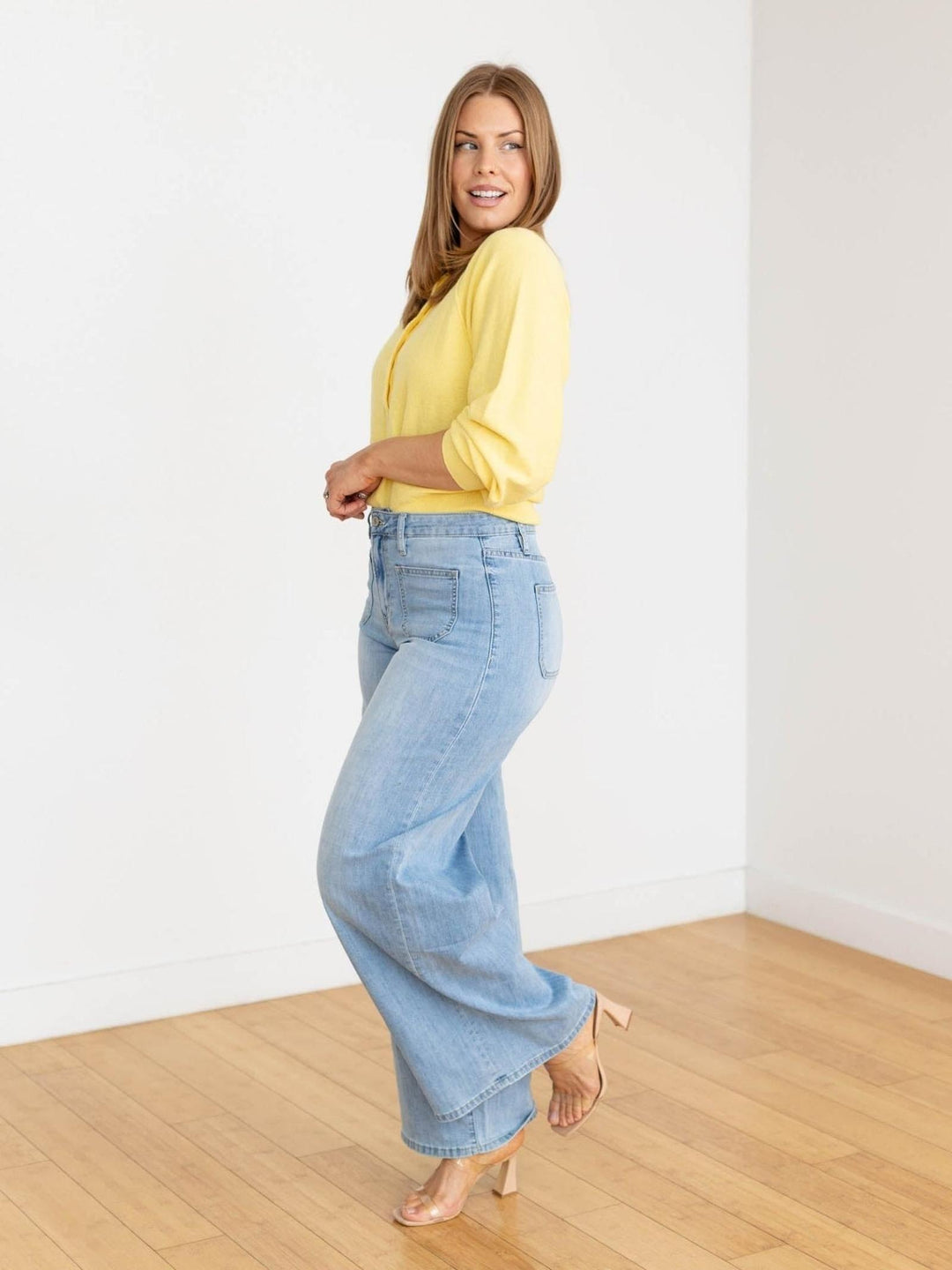Woman wearing a yellow top and light blue jeans standing in a room with wooden flooring and white walls.