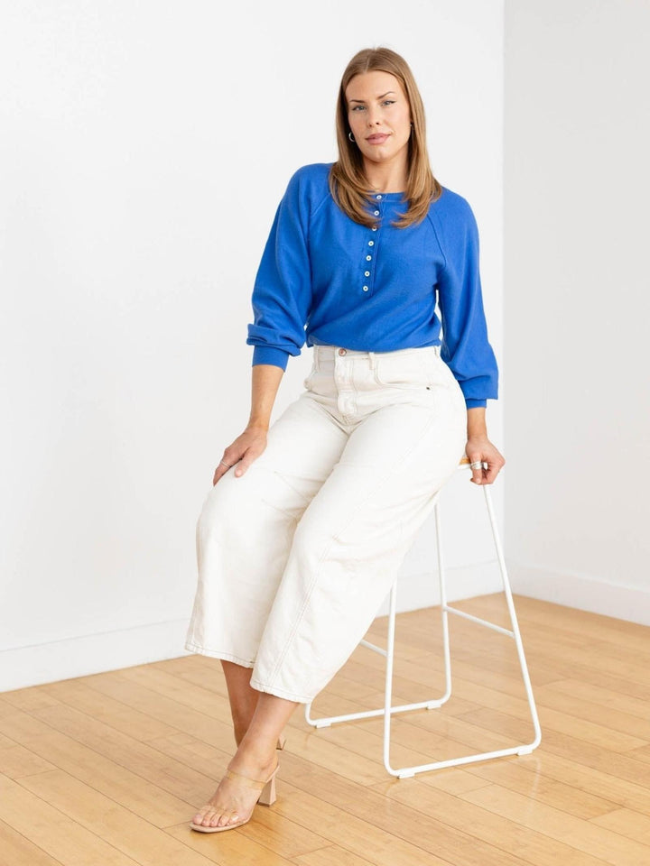 Woman wearing a blue top and white pants standing next to a white stool on a wooden floor.