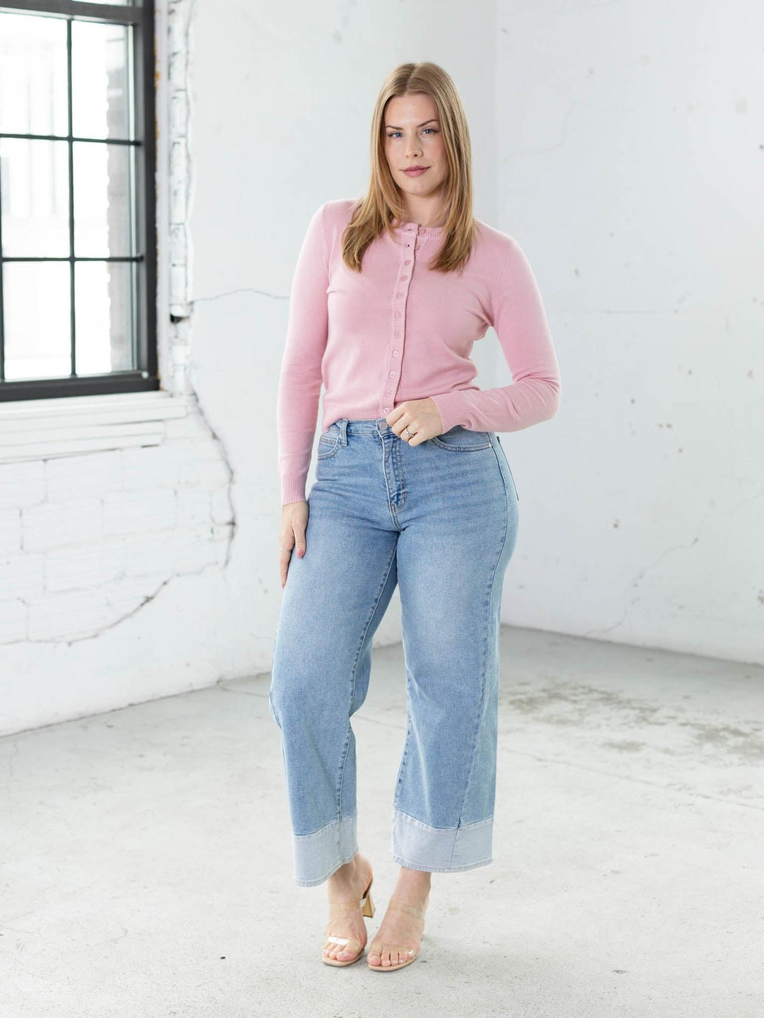 Woman wearing a pink cardigan and light blue jeans in a white room.
