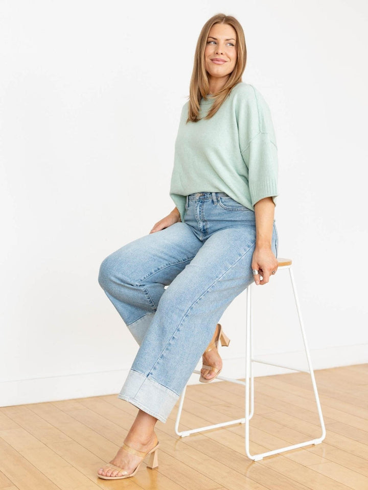 Woman sitting on a white stool wearing a light green top and light blue jeans against a white background.