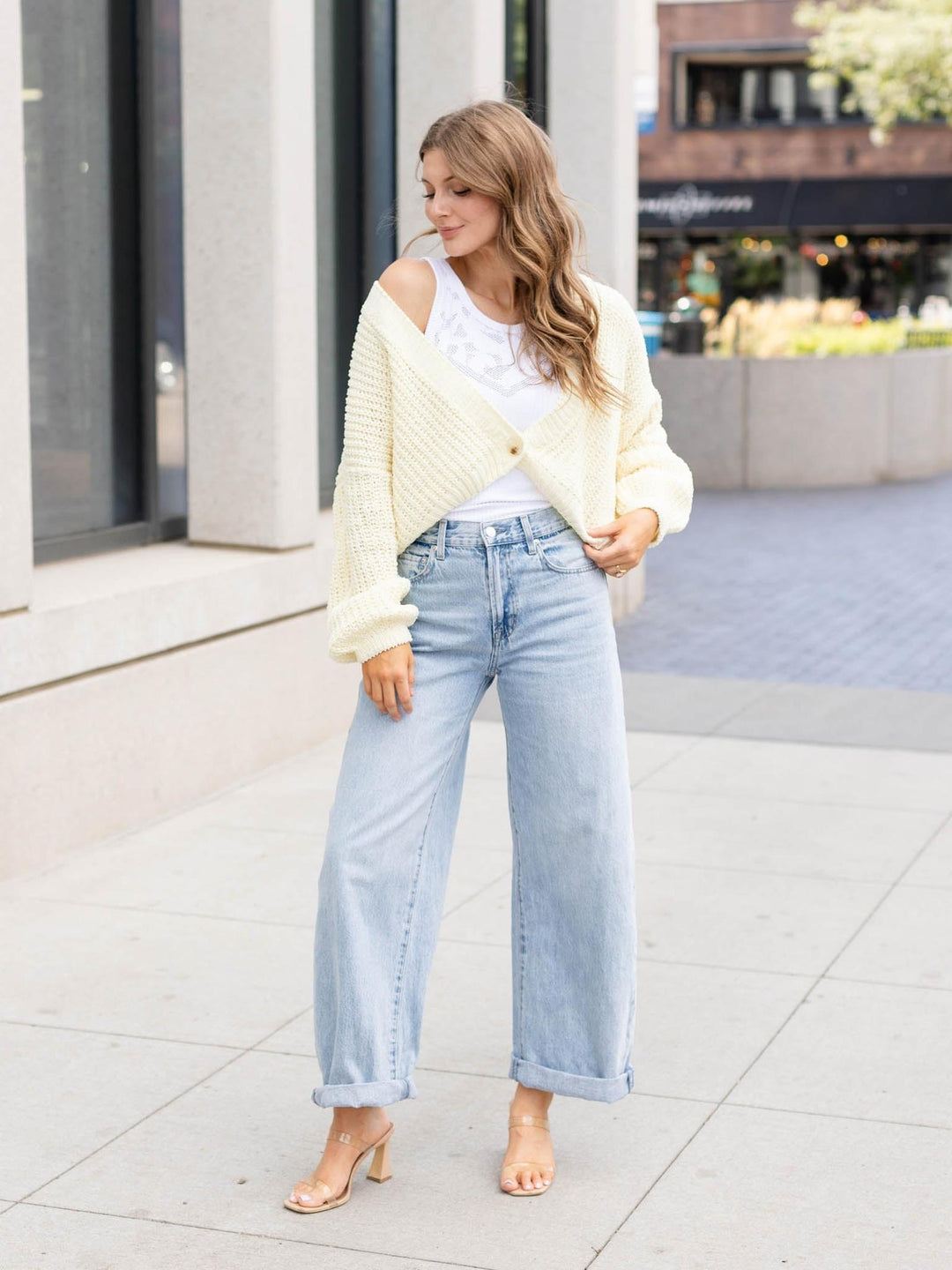 A woman stands on a city sidewalk wearing a cream ribbed knit cardigan over a white top, light blue wide-leg jeans, and clear high-heeled sandals.