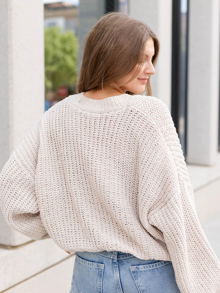 A woman wearing a cream-colored, chunky open-knit sweater and light blue jeans, photographed from behind in an outdoor urban setting.