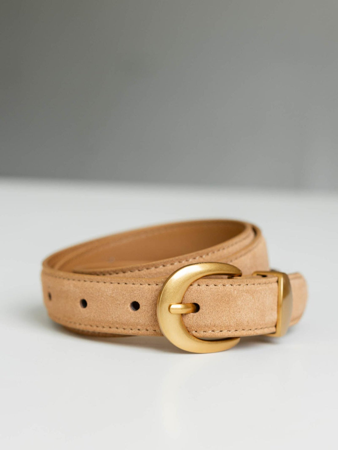 Tan suede belt with a curved gold buckle, coiled on a white surface.