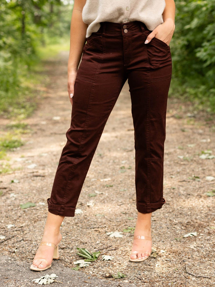A person wearing dark brown cropped pants with rolled cuffs, standing on a dirt path outdoors with green foliage in the background.