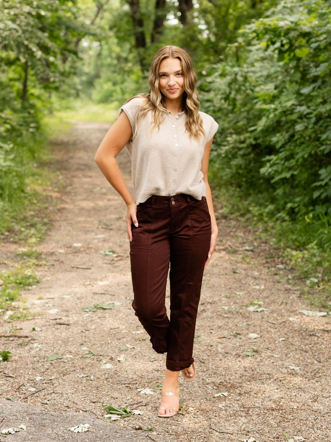 A woman walking on a dirt path in a wooded area, wearing a light beige short-sleeve button-up shirt and dark brown pants.