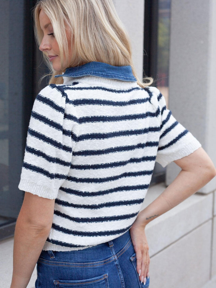 A woman wearing a short-sleeve knit top with navy blue and white horizontal stripes, paired with blue jeans, shown from a side-back angle in an outdoor setting.