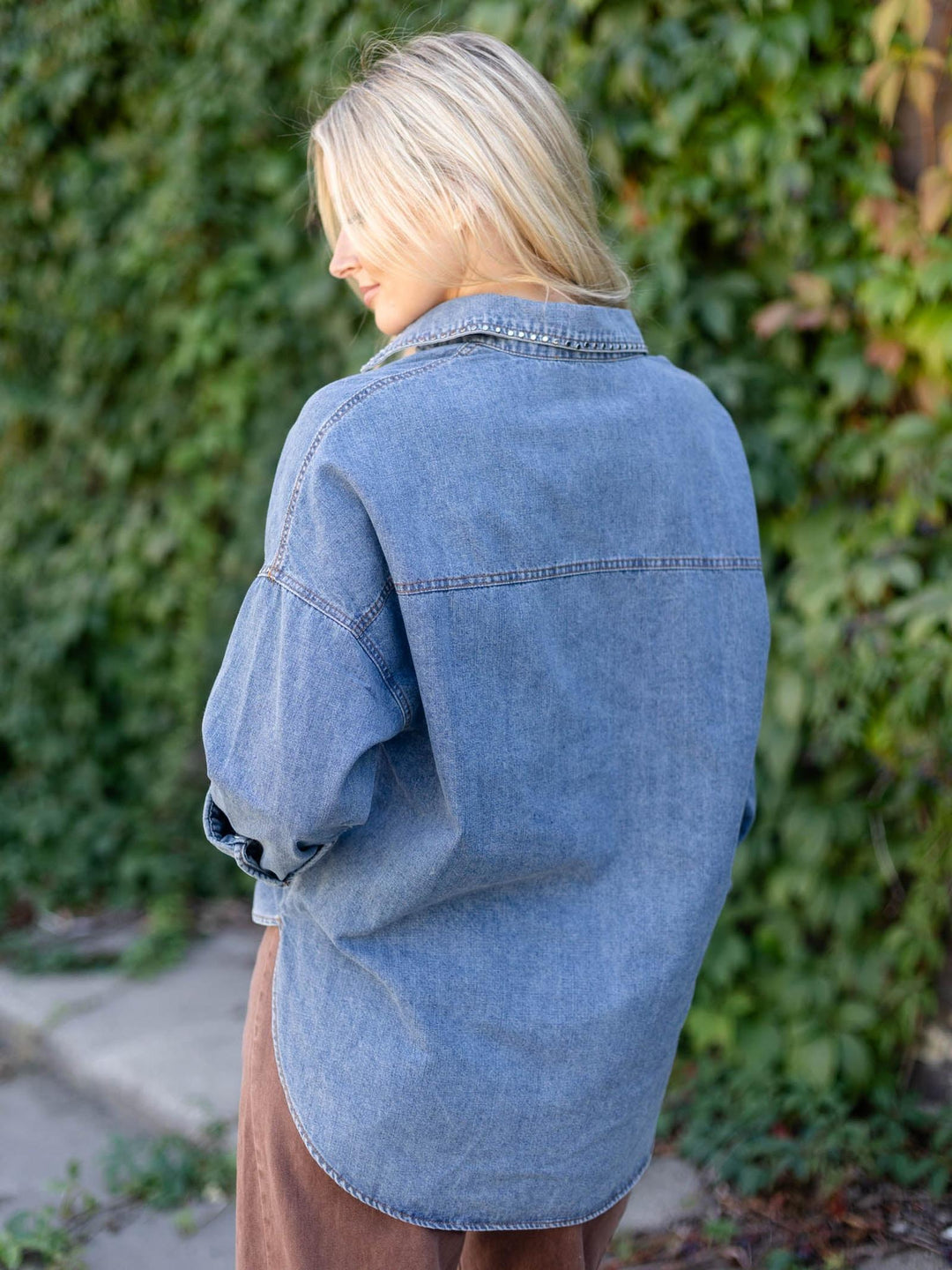 A woman with blonde hair is shown from behind wearing an oversized light blue denim shirt, standing outdoors against a background of green ivy.
