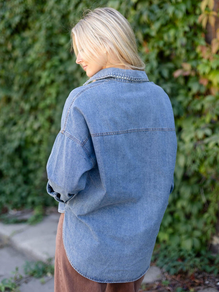 A woman with blonde hair is shown from behind wearing an oversized light blue denim shirt, standing outdoors against a background of green ivy.
