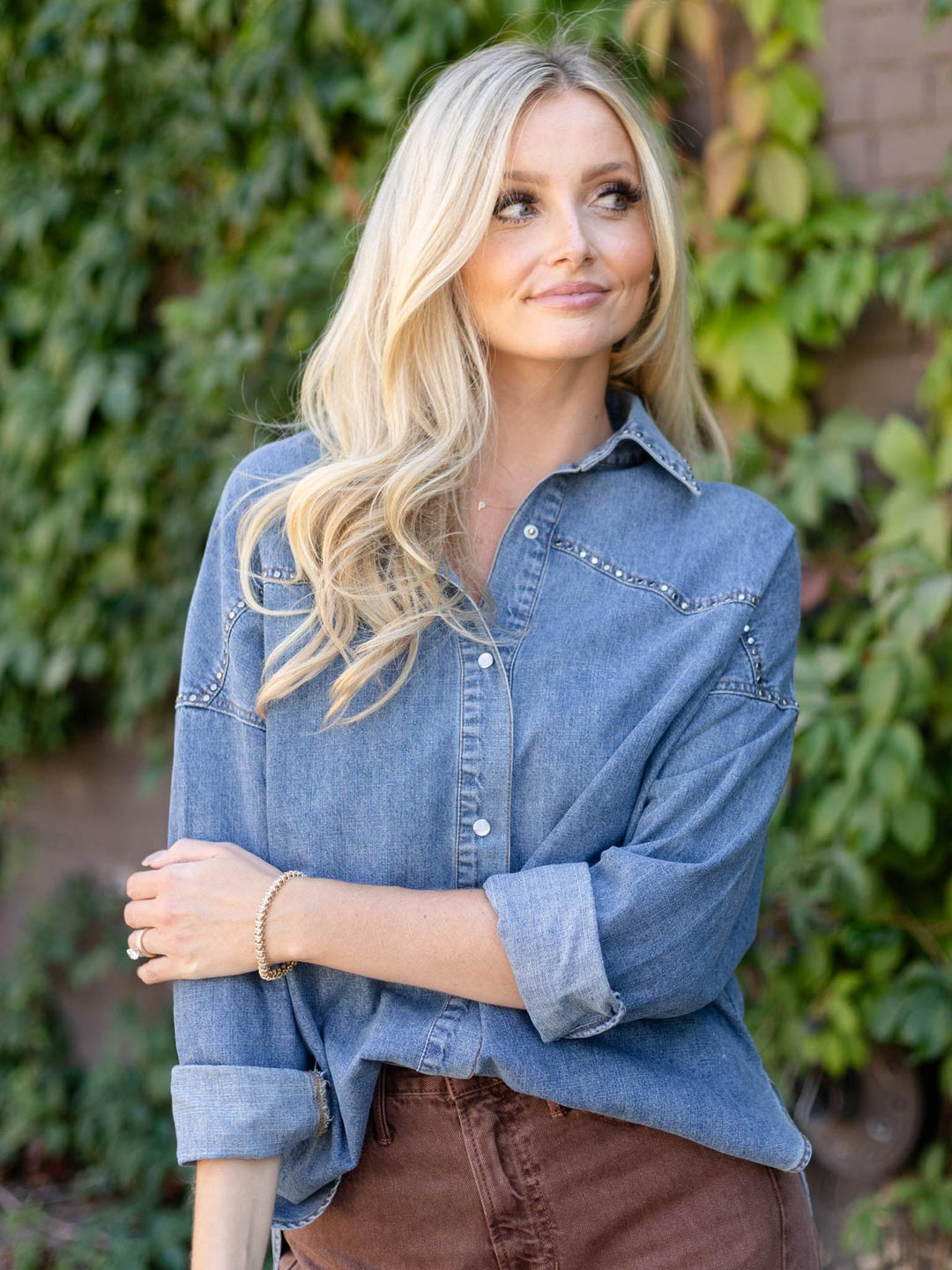 A blonde woman wearing a blue denim button-up shirt with western-style stitching and brown pants, standing outdoors in front of a green leafy background.