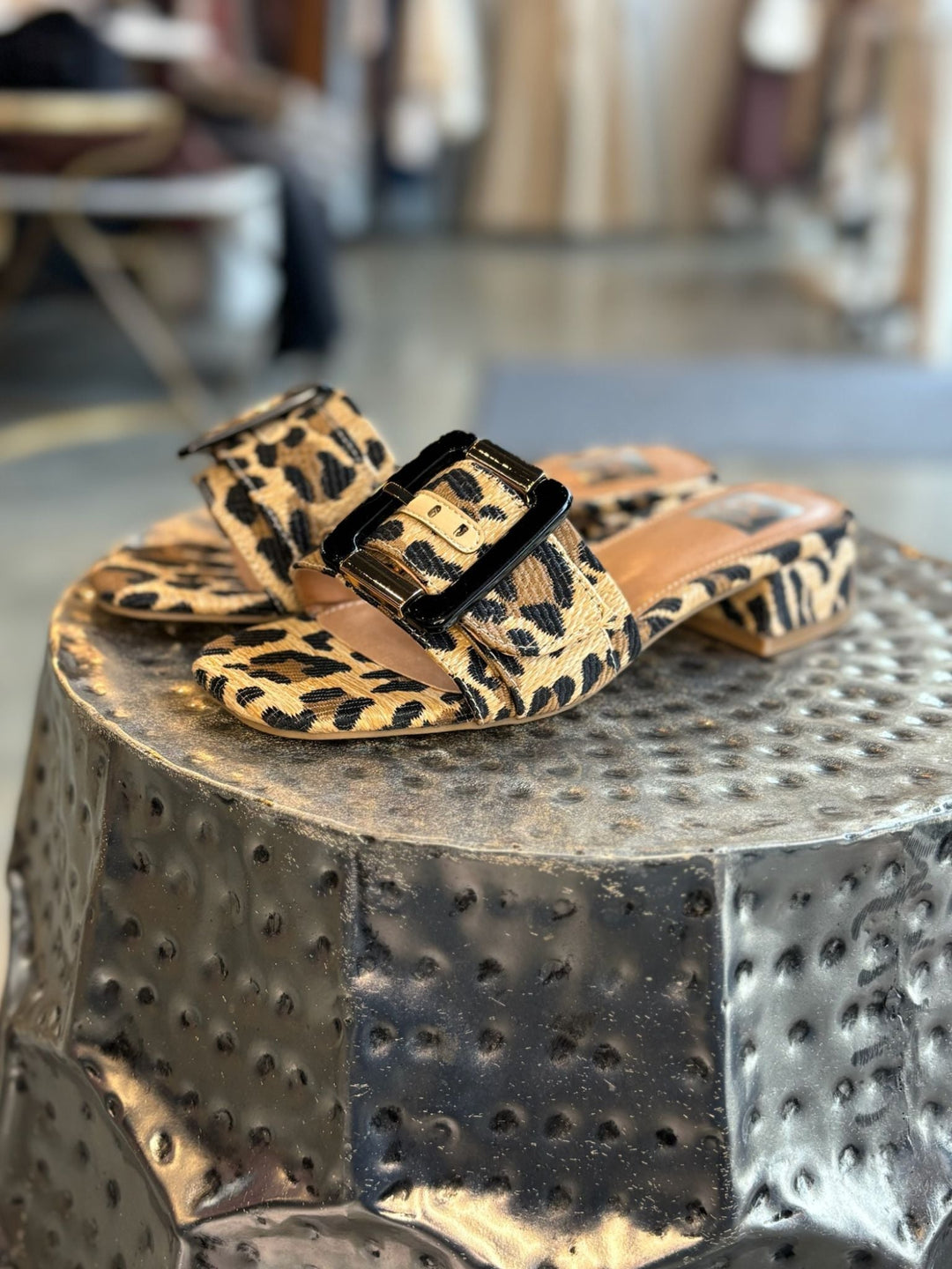 Leopard print sandals with a black buckle on a textured metallic surface.