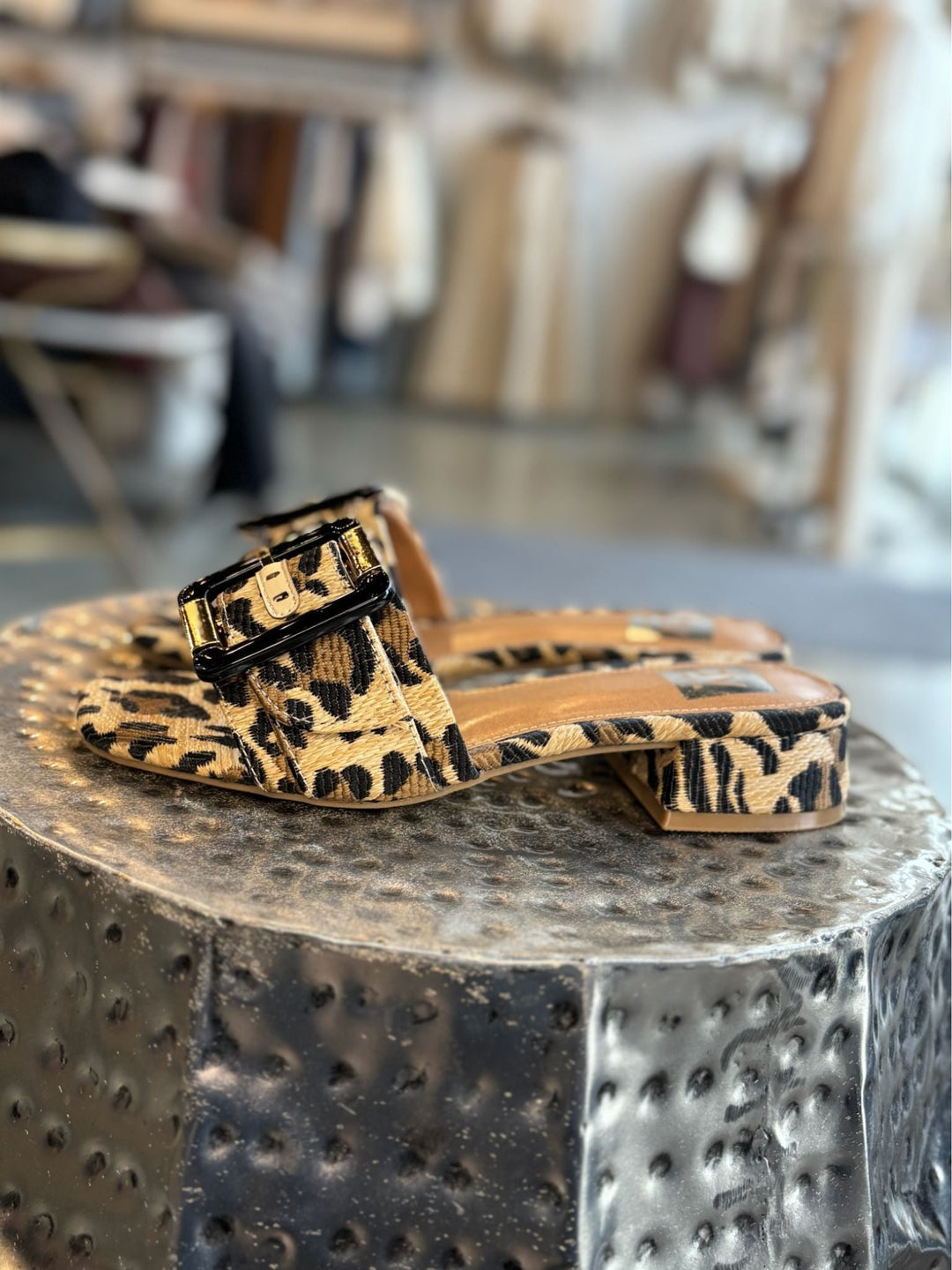 Leopard print sandals with a black buckle on a textured metallic surface.