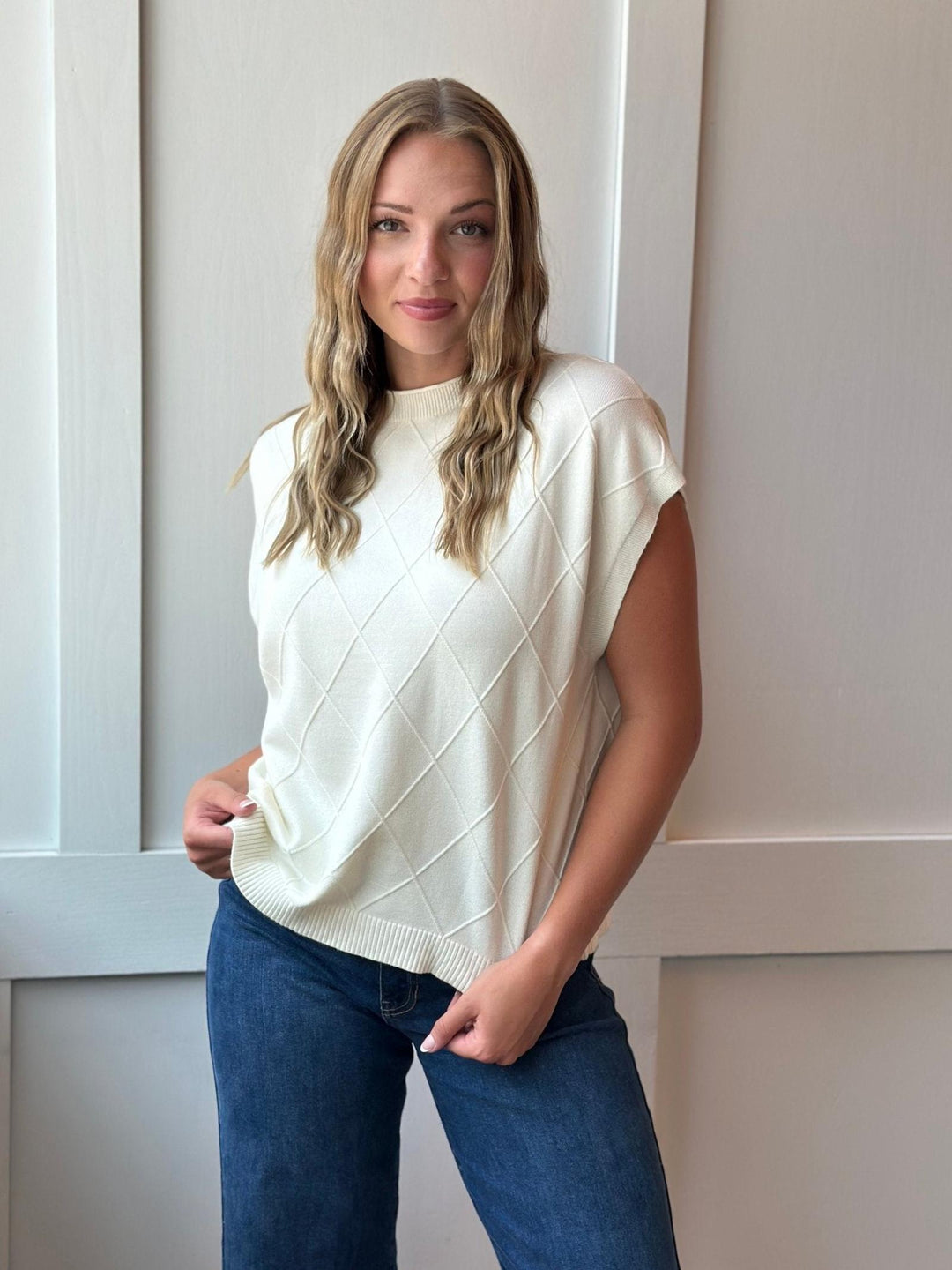A woman wearing a cream-colored sleeveless knit sweater with a diamond pattern, paired with blue jeans, standing against a white paneled wall.