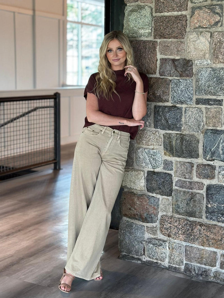 A woman stands indoors leaning against a stone wall, wearing a dark burgundy short-sleeve top and light beige wide-leg pants with clear high-heel sandals.