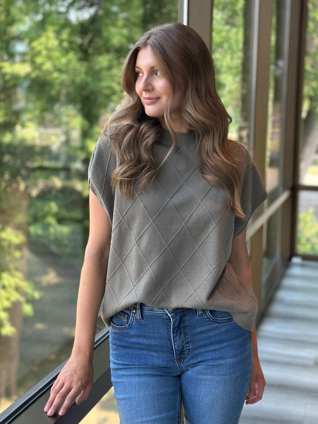 A woman wearing a short-sleeve knit top with a textured argyle pattern in taupe-gray, paired with blue jeans, standing on a balcony with green trees visible in the background.