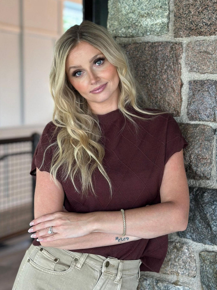 A blonde woman wearing a short-sleeve maroon knit top and light beige pants, leaning against a stone wall.
