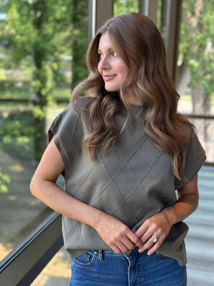 A woman with long brown hair wearing a muted olive green short-sleeve top with a diamond quilted pattern, paired with blue jeans, standing in front of a glass window with a blurred outdoor background.