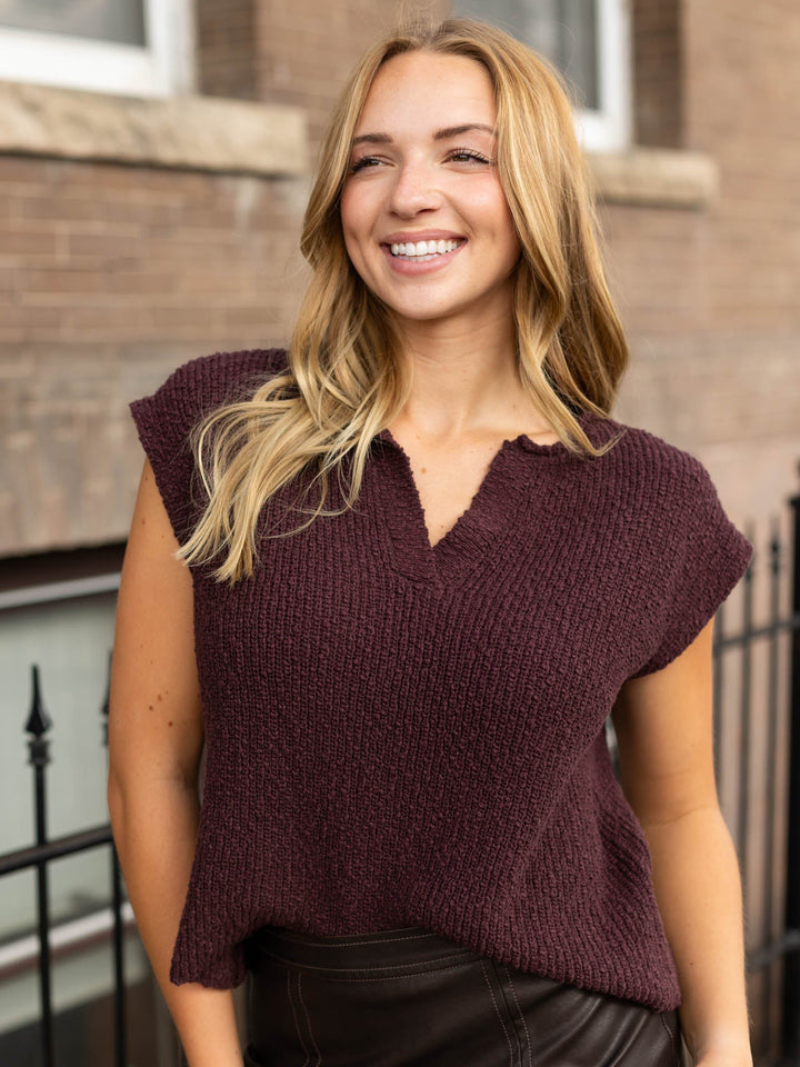 A smiling blonde woman wearing a textured, short-sleeve knit top in a deep plum-burgundy color, standing outdoors in front of a brick building.