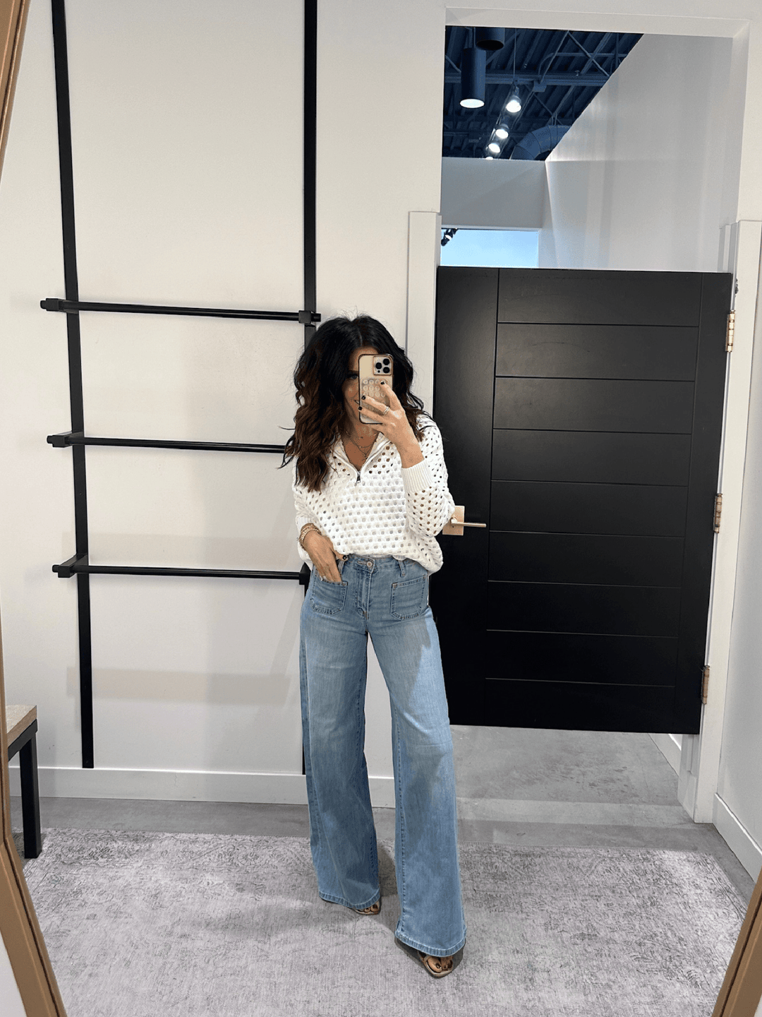 Person taking a mirror selfie wearing a polka dot blouse and blue jeans in a modern interior setting.