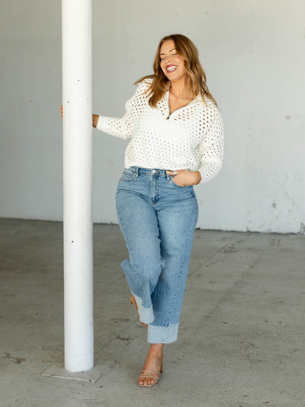 Woman wearing a white blouse and blue jeans standing against a white wall.