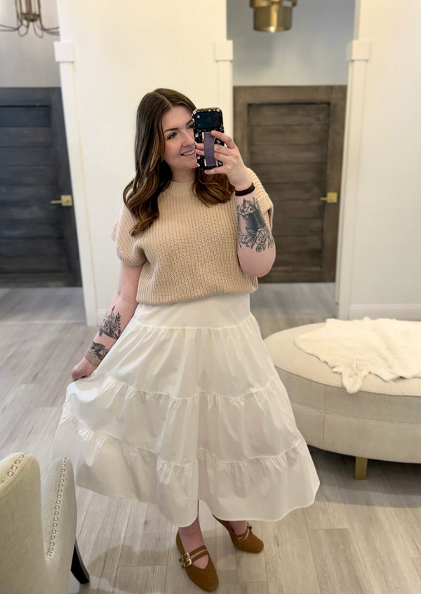 Woman taking a mirror selfie wearing a beige sweater and white skirt in a room.