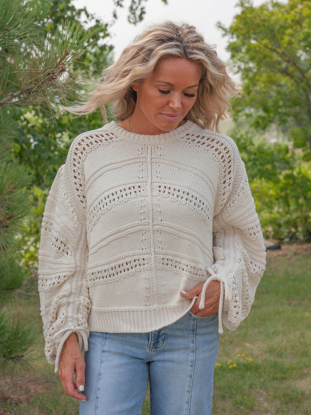 A woman wearing a cream-colored, textured open-knit sweater with balloon sleeves and light blue jeans, standing outdoors in a grassy area with trees in the background.