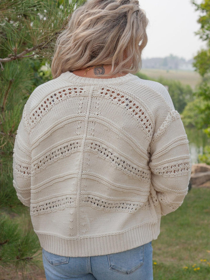 A woman viewed from behind wearing a cream-colored, textured knit sweater with horizontal openwork patterns, paired with light blue jeans in an outdoor setting.