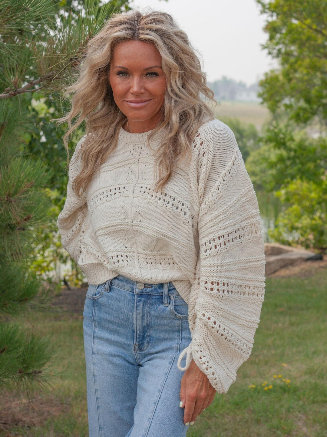 A woman wearing a cream-colored, textured knit sweater with wide sleeves and blue jeans, standing outdoors in a grassy area with trees in the background.