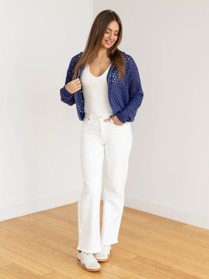 Woman wearing a blue cardigan, white top, and white pants in a minimalistic room.
