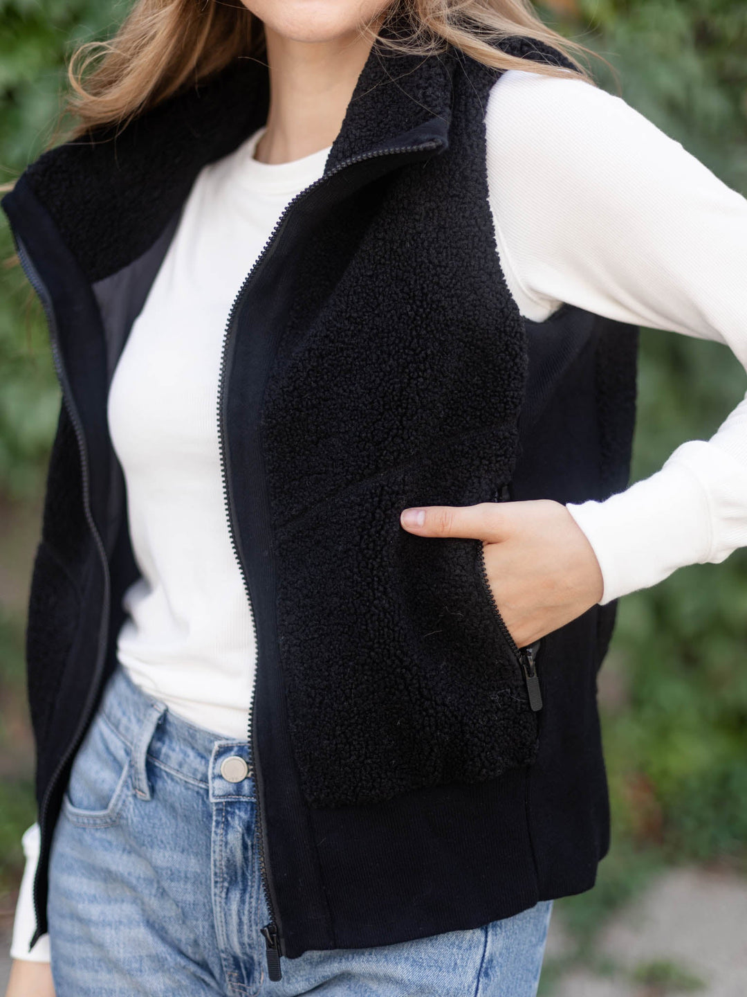 A woman wearing a black fleece zip-front vest over a white long-sleeve top and blue jeans, photographed outdoors with a blurred green background.