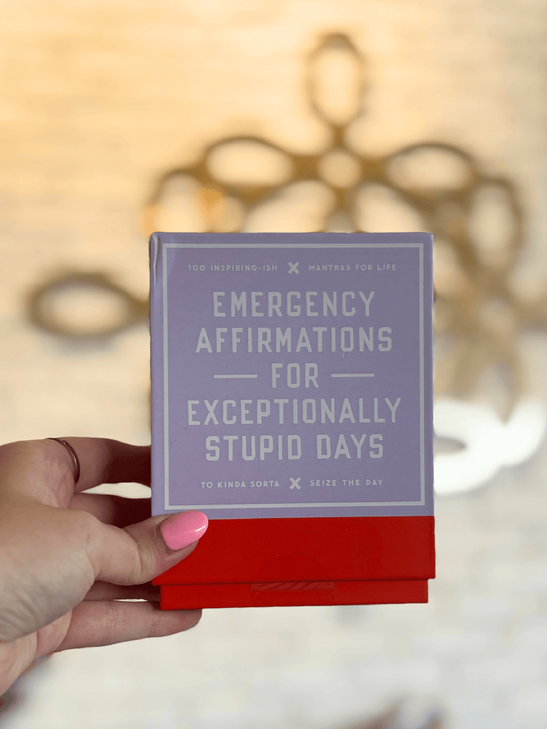 A hand with pink nail polish holds a small square card or box featuring the text &