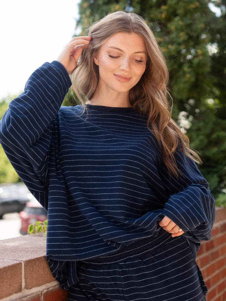 A woman wearing a navy blue long-sleeve top with thin white vertical stripes, standing outdoors against a blurred background of green trees and a brick wall.