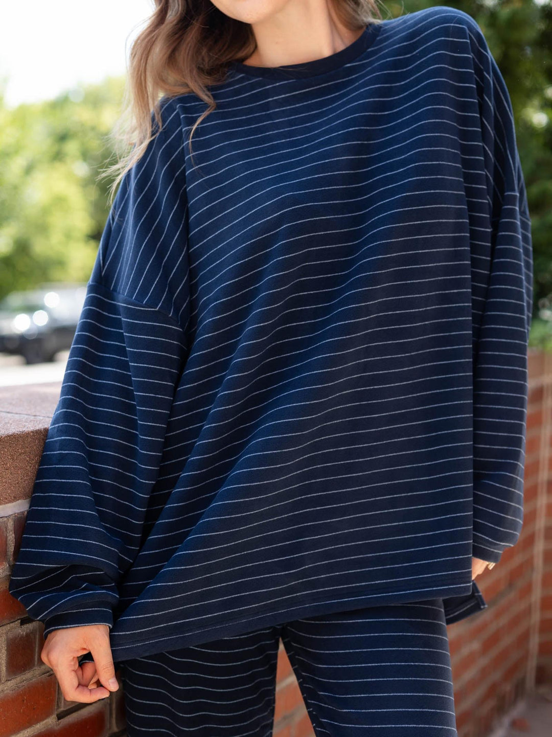 A person wearing an oversized long-sleeve top and matching pants in dark navy blue with thin white horizontal stripes, standing outdoors against a brick wall and blurred greenery.