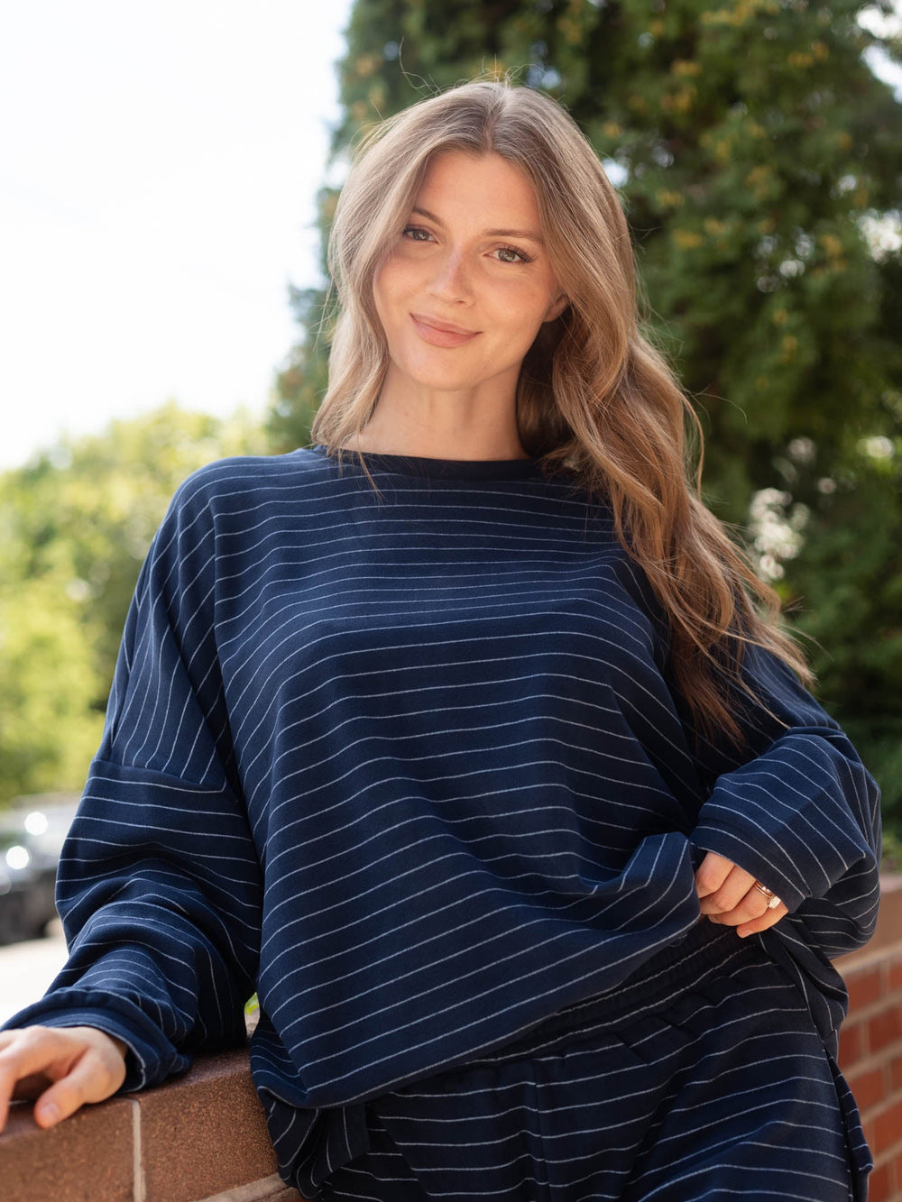 A woman with long blonde hair wearing a navy blue long-sleeve top with thin white vertical stripes, leaning against a brick wall outdoors with green trees in the background.