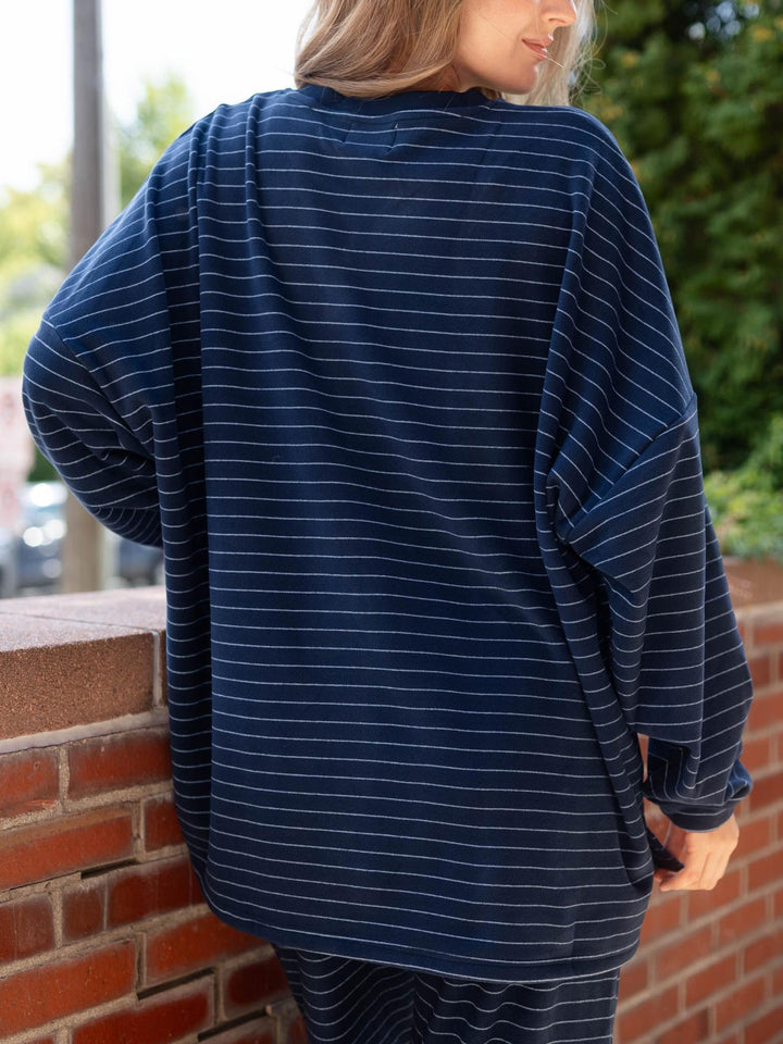 A woman wearing an oversized navy blue long-sleeve top with thin white horizontal stripes, shown from the back while leaning against a brick wall outdoors.