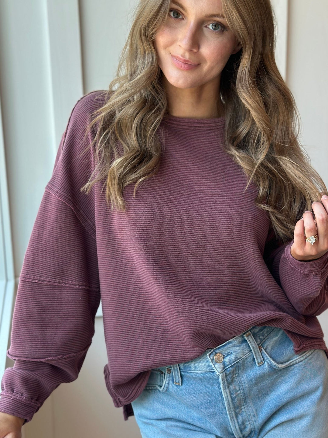 A woman wearing a ribbed, long-sleeve crewneck sweatshirt in a dusty mauve-purple color, paired with light-wash blue jeans.