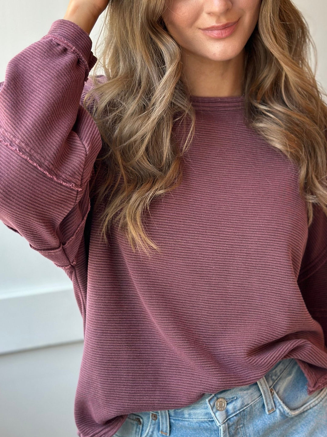 A woman wearing a ribbed long-sleeve crewneck top in a dusty mauve or plum color, paired with light blue jeans.
