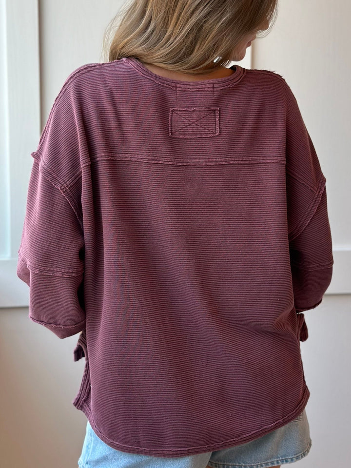A person shown from the back wearing an oversized ribbed knit top in a muted dusty mauve or burgundy tone, paired with light blue denim bottoms, against a plain indoor wall.