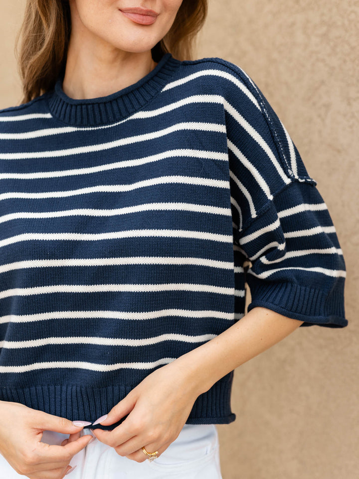 A woman wearing a navy blue and white horizontally striped knit sweater with three-quarter sleeves, paired with white pants, set against a neutral textured beige wall.