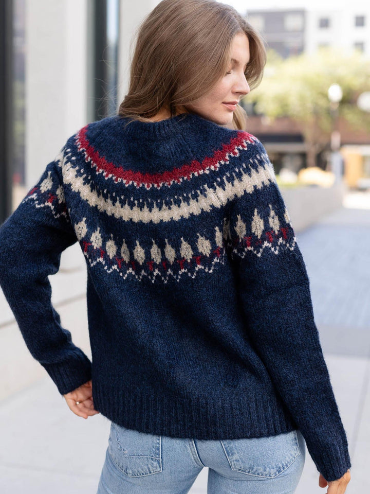 A woman wearing a navy blue knit sweater with a traditional Fair Isle pattern in cream and red, shown from the back in an outdoor urban setting.